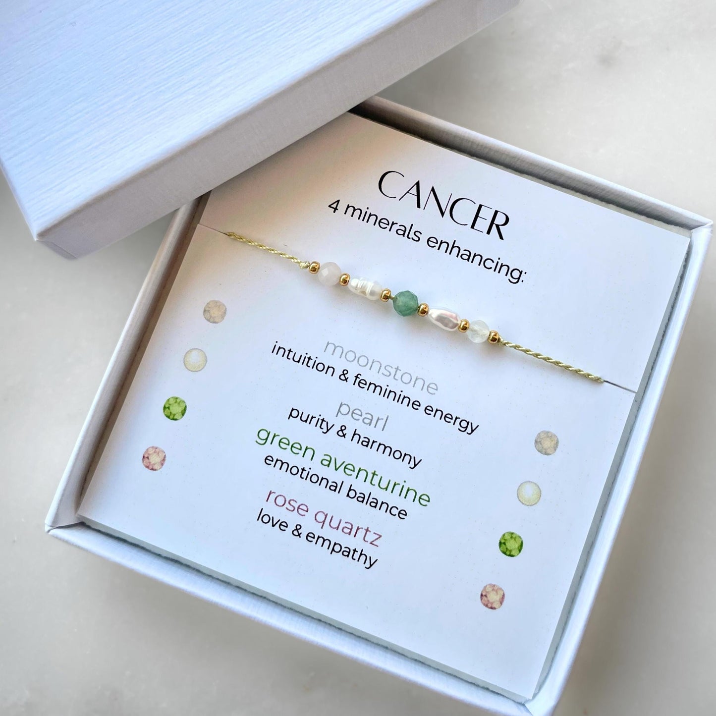 White gift box that contains a mineral bracelet for a Cancer Zodiac sign. The bracelet is displayed on a card that describes the minerals and their meanings. There are five minerals on the bracelet: moonstone, two pearls, green aventurine and rose quartz