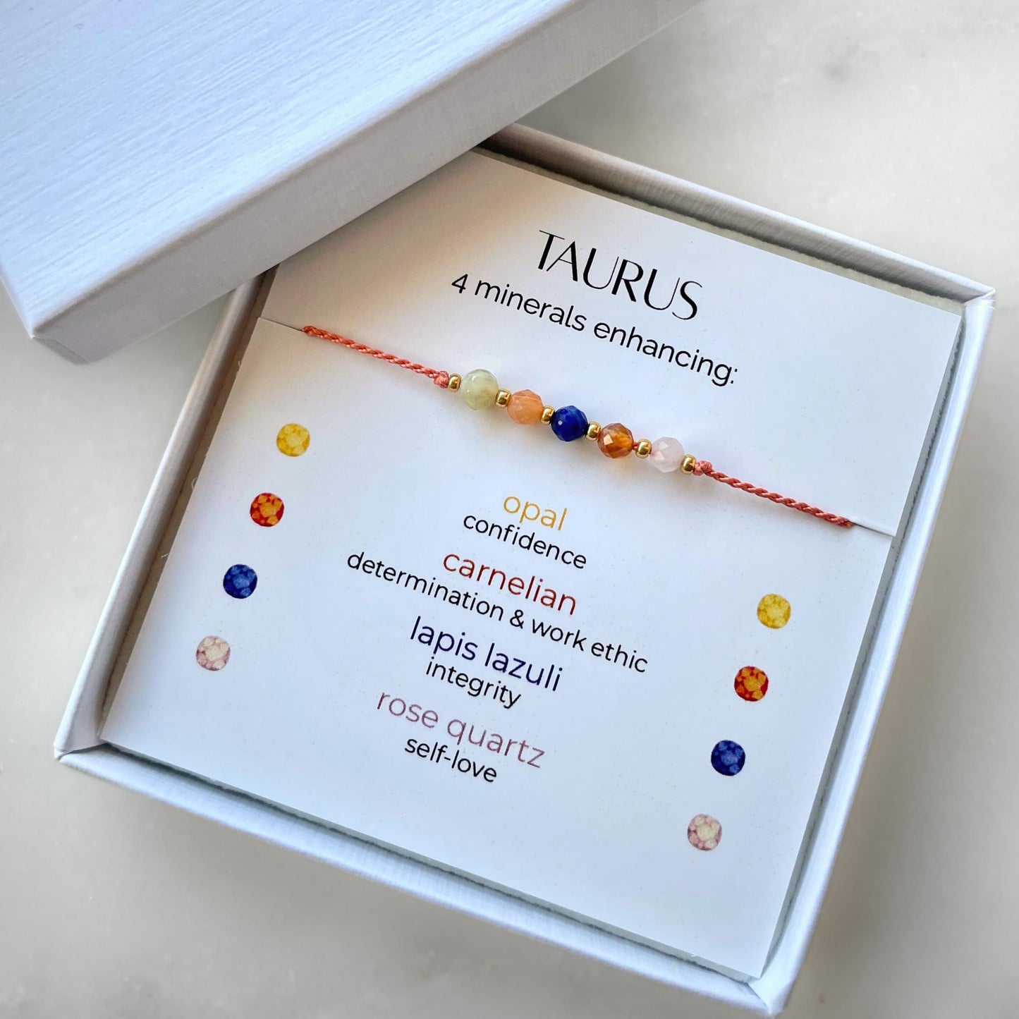 White gift box that contains a mineral bracelet for a Taurus Zodiac sign. The bracelet is displayed on a card that describes the minerals and their meanings. There are five minerals on the bracelet: opal, two carnelians, lapis lazuli and rose quartz