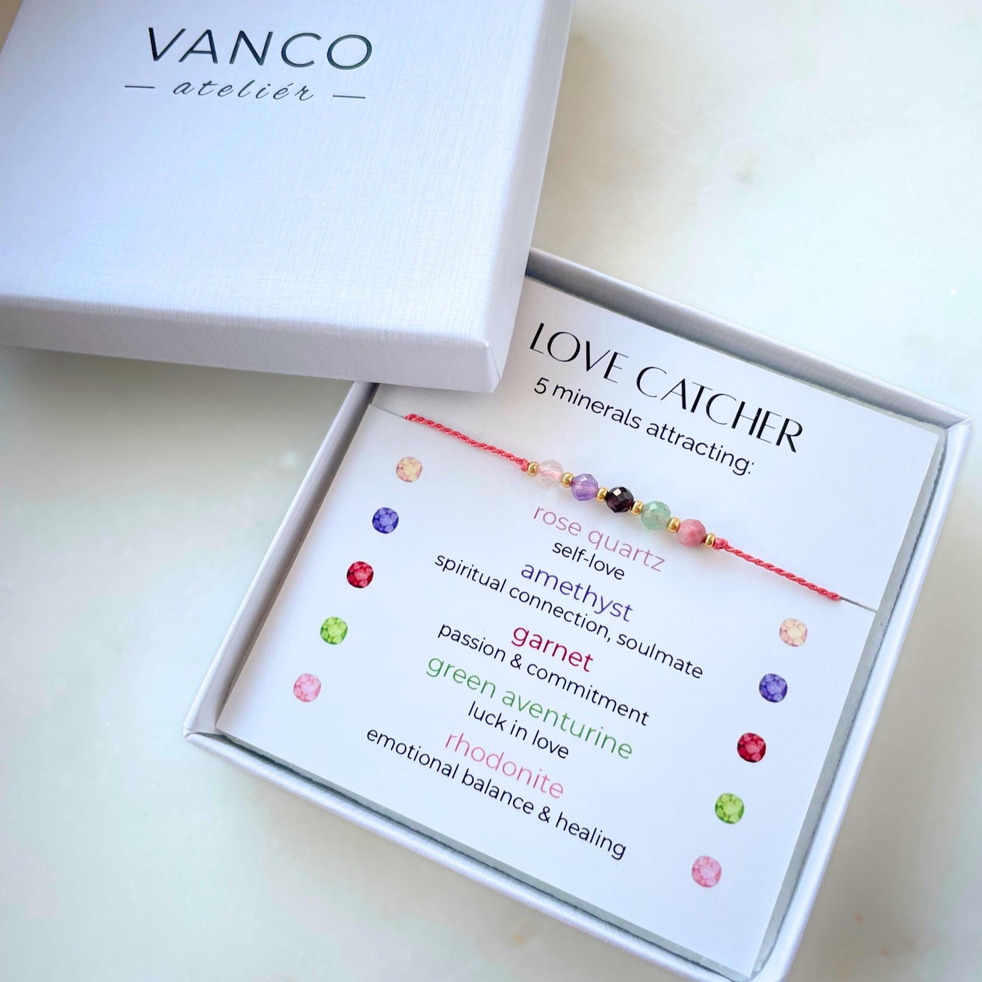 A white gift box that contains a bracelet made of pink silk string with five mineral beads, put on a promotional card. The bracelet's theme is Love, the name of the bracelet is Love Catcher which is written in the promotional card. The card also explains each stone and what each stone represents: Rose Quartz for Self-love, Amethyst for Spiritual Connection & Soulmate, Garnet for Passion & Commitment, Green Aventurine for Luck in Love, Rhodonite for Emotional Balance & Healing