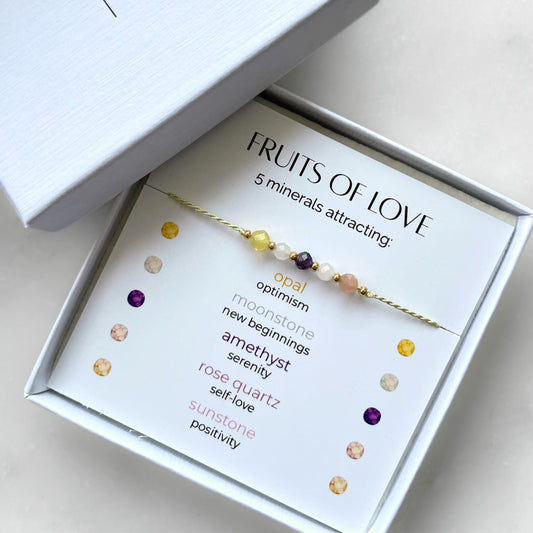 A white gift box that contains a bracelet made of green silk string with five mineral beads, put on a promotional card. The bracelet's theme is Fertility, the name of the bracelet is Fruits of Love which is written in the promotional card. The card also explains each stone and what each stone represents: Opal for Optimism, Moonstone for New Beginnings, Amethyst for Serenity, Rose Quartz for Self-love, Sunstone for Positivity