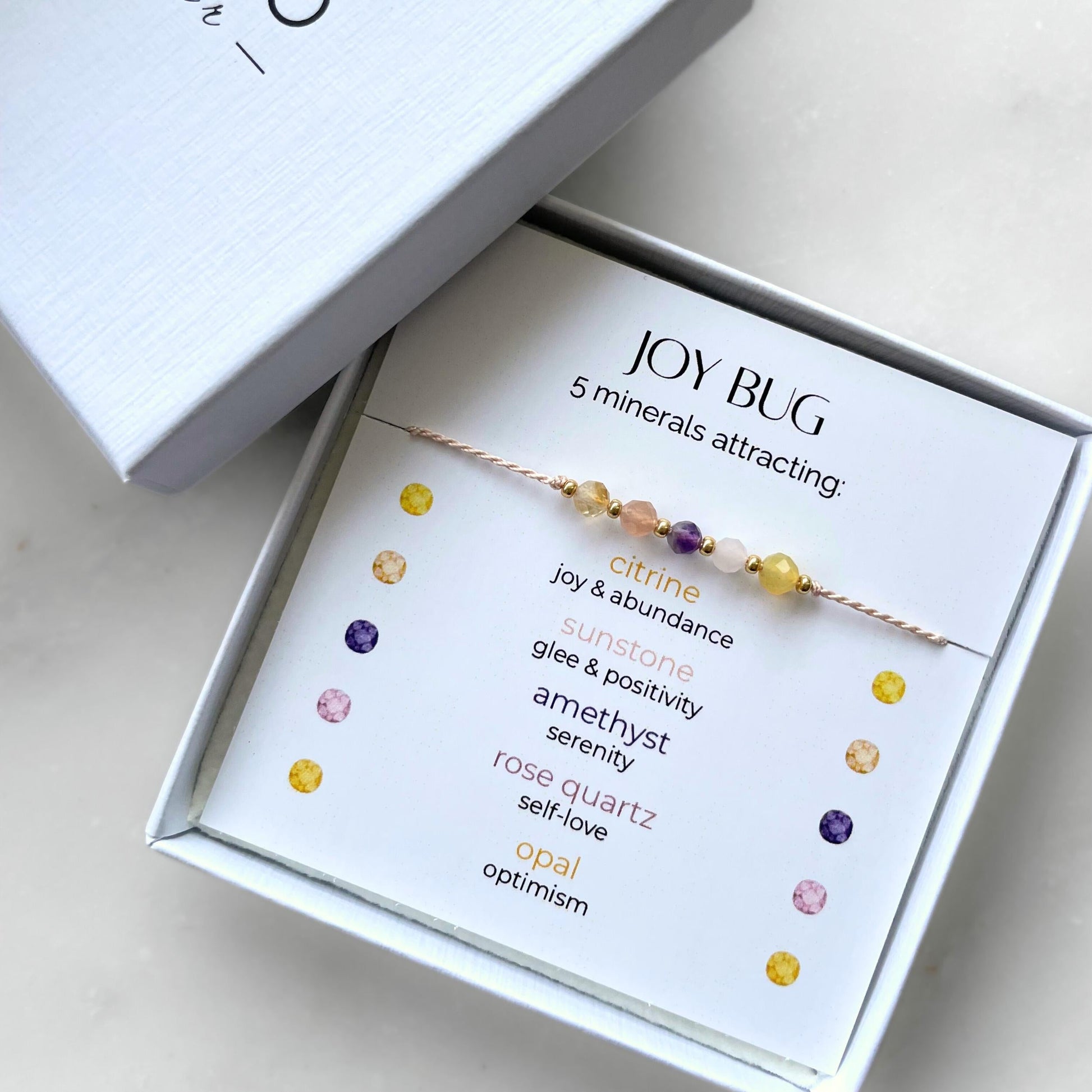 A white gift box that contains a bracelet made of pink silk string with five mineral beads, put on a promotional card. The bracelet's theme is Happiness, the name of the bracelet is Joy Bug which is written in the promotional card. The card also explains each stone and what each stone represents: Citrine for Joy & Abundance, Sunstone for Glee & Positivity, Amethyst for Serenity, Rose Quartz for Self-love, Opal for Optimism