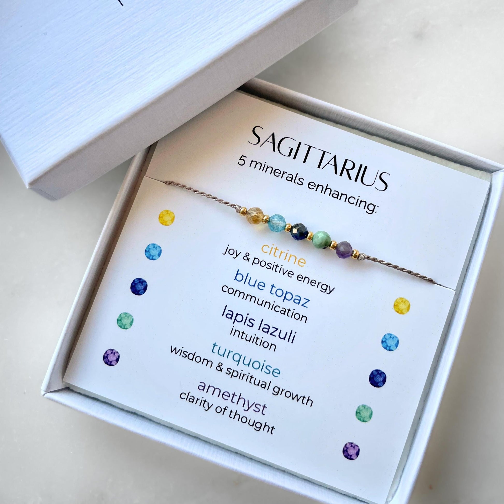 A white gift box that contains a bracelet made of grey silk string with five mineral beads, put on a promotional card. The name of the bracelet is Sagittarius which is written in the promotional card. The card also explains each stone and what each stone represents: Citrine for Joy and Positive Energy, Blue Topaz for Communication, Lapis Lazuli for Intuition, Turquoise for Wisdom and Spiritual Growth, Amethyst for Clarity of Thought