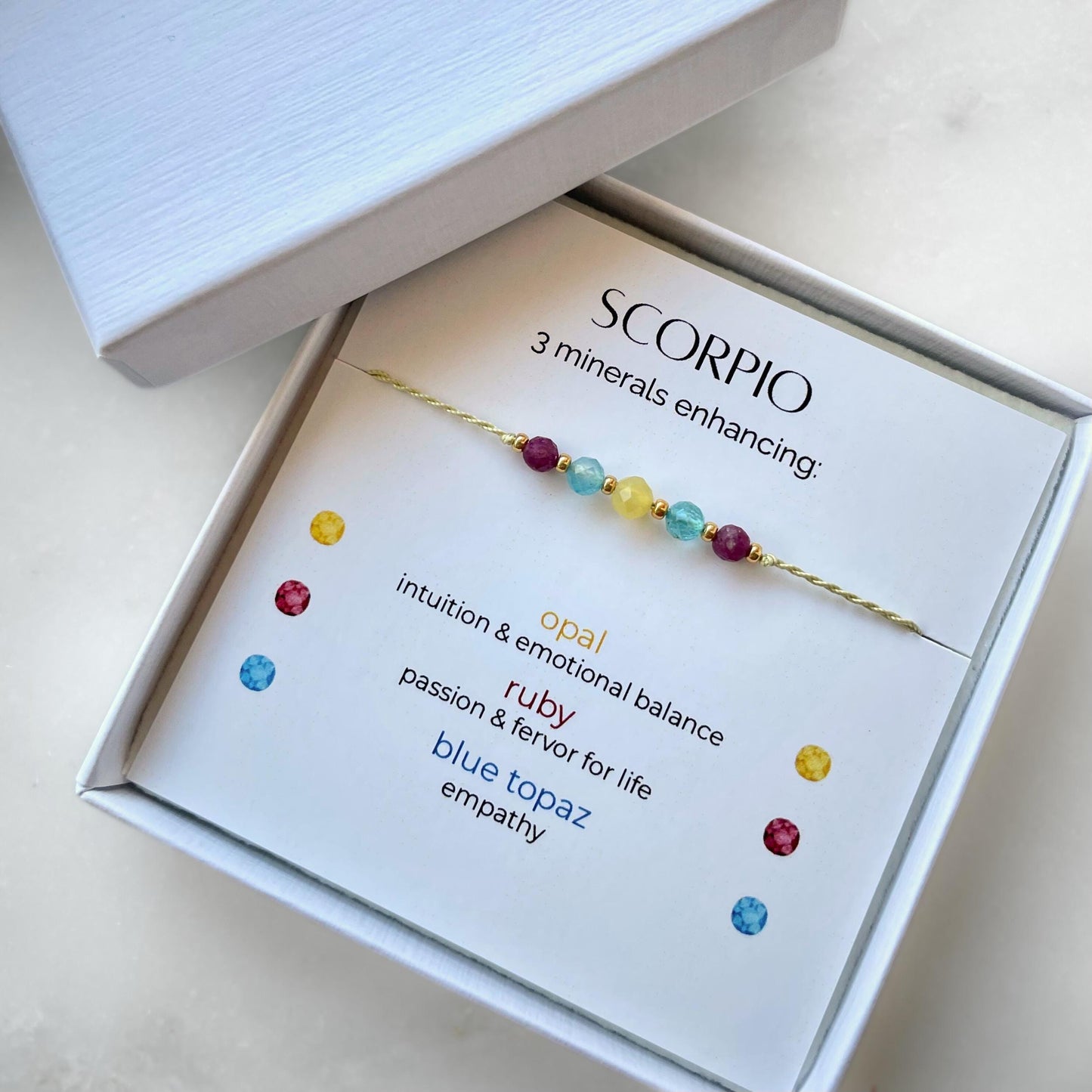 White gift box that contains a mineral bracelet for a Scorpio Zodiac sign. The bracelet is displayed on a card that describes the minerals and their meanings. There are five minerals on the bracelet: Two rubies, two blue topazes and one opal.
