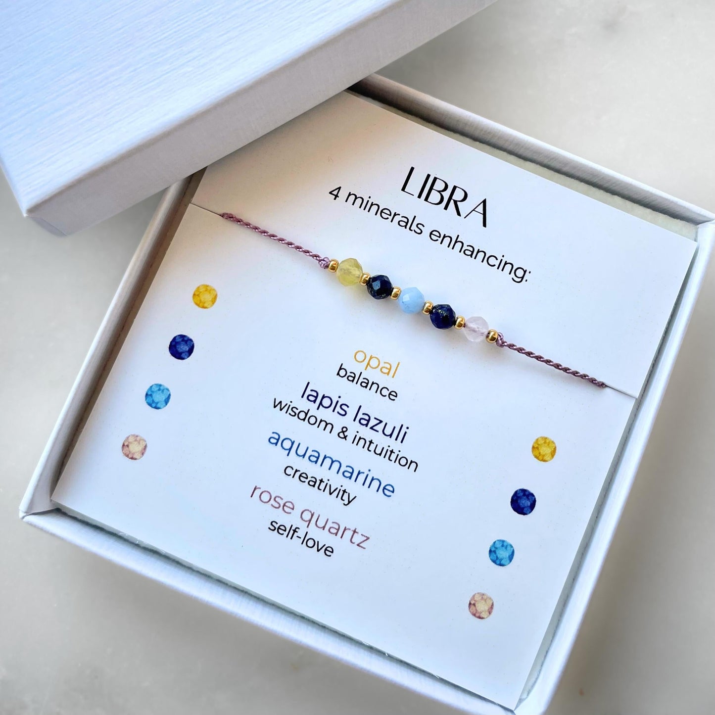 White gift box that contains a mineral bracelet for a Libra Zodiac sign. The bracelet is displayed on a card that describes the minerals and their meanings. There are five minerals on the bracelet: Opal, two Lapis Lazulis, Aquamarine and Rose Quartz