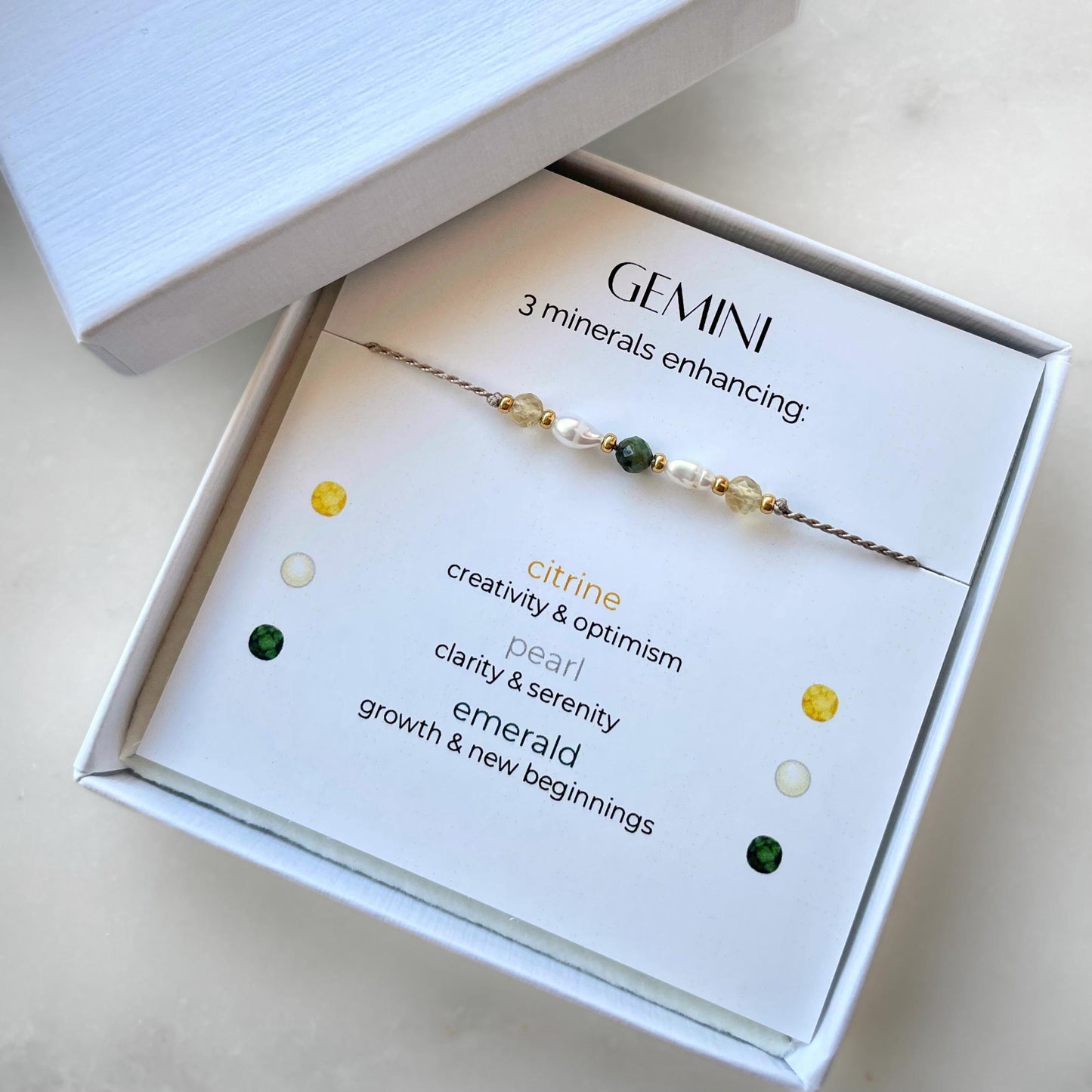 White gift box that contains a mineral bracelet for a Gemini Zodiac sign. The bracelet is displayed on a card that describes the minerals and their meanings. There are five minerals on the bracelet: two citrines, two pearls and one emerald