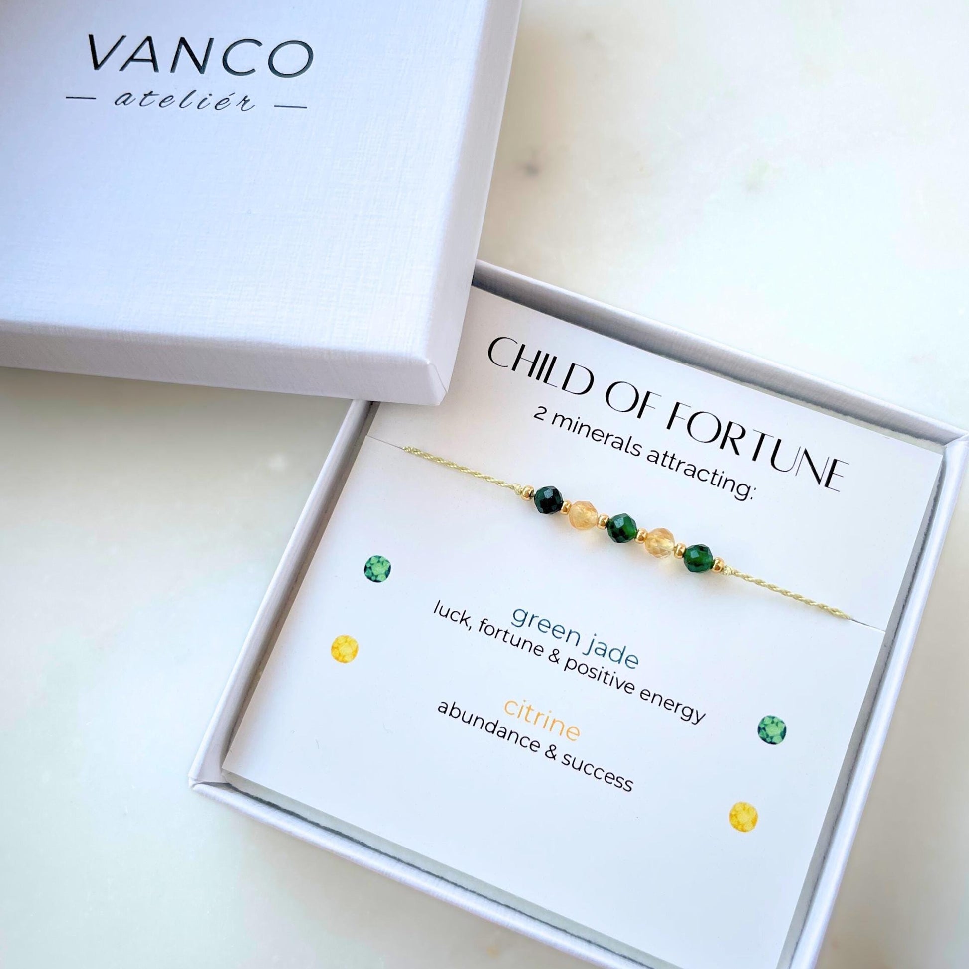 A white gift box that contains a bracelet made of green silk string with five mineral beads, put on a promotional card. The bracelet's theme is Luck, the name of the bracelet is Child of Fortune which is written in the promotional card. The card also explains each stone and what each stone represents: Green Jade for Luck, Fortune & Positive Energy, Citrine for Abundance & Success