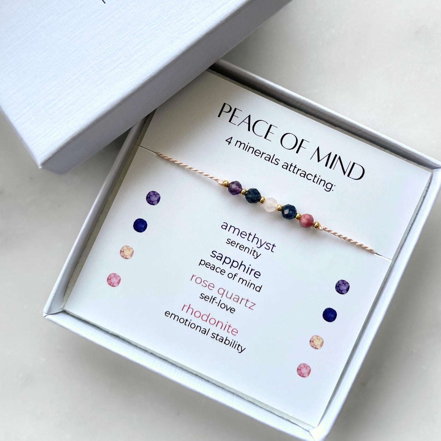 A white gift box that contains a bracelet made of pink silk string with five mineral beads, put on a promotional card. The bracelet's theme is Serenity, the name of the bracelet is Peace of Mind which is written in the promotional card. The card also explains each stone and what each stone represents: Amethyst for Serenity, Sapphire for Peace of Mind, Rose Quartz for Self-love, Rhodonite for Emotional Stability
