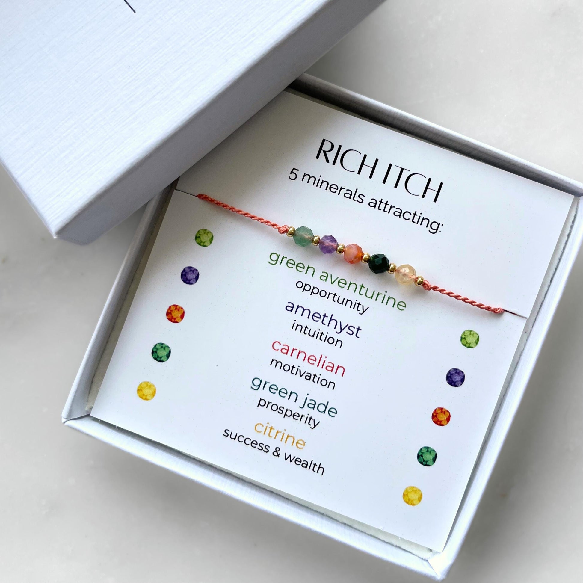 A white gift box that contains a bracelet made of tangerine silk string with five mineral beads, put on a promotional card. The bracelet's theme is Wealth, the name of the bracelet is Rich Itch which is written in the promotional card. The card also explains each stone and what each stone represents: Green Aventurine for Opportunity, Amethyst for Intuition, Carnelian for Motivation, Green Jade for Prosperity, Citrine for Success & Wealth