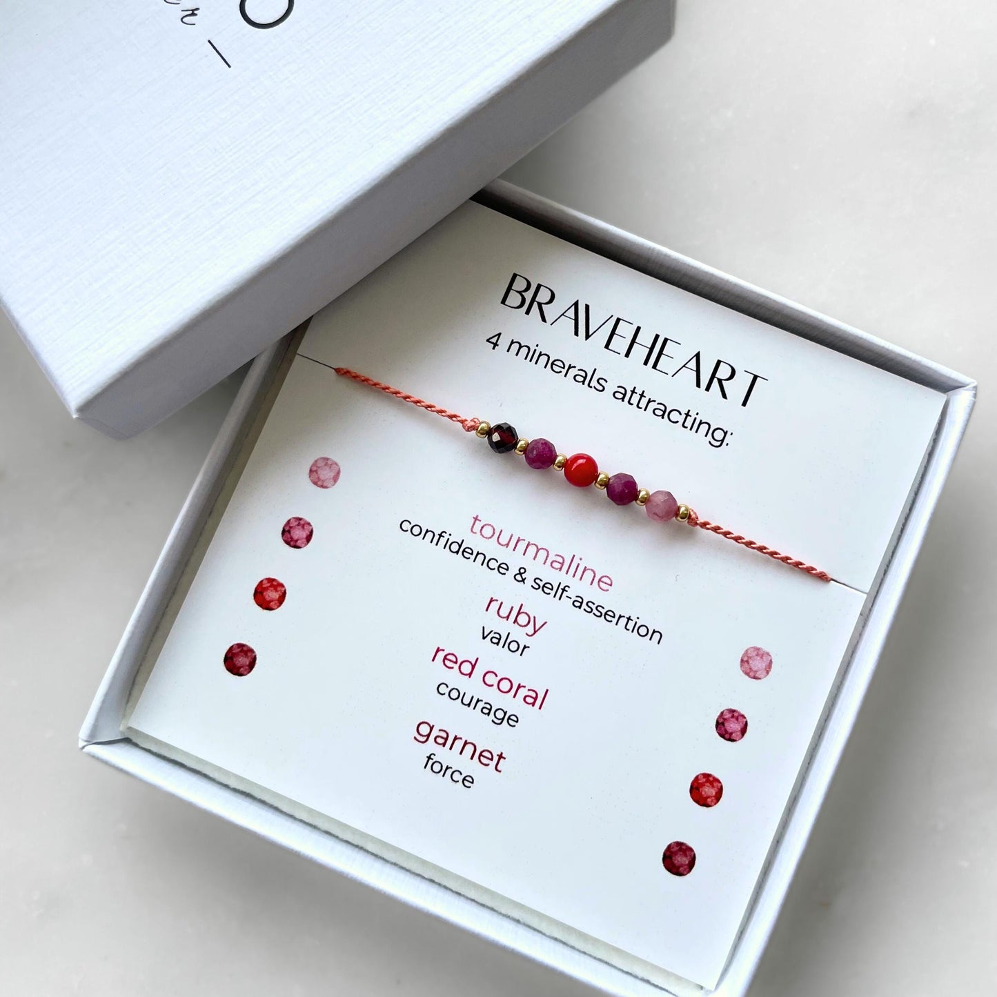A white gift box that contains a bracelet made of tangerine silk string with five mineral beads, put on a promotional card. The bracelet's theme is Courage, the name of the bracelet is Braveheart which is written in the promotional card. The card also explains each stone and what each stone represents: Red Coral for Courage, Ruby for Valor, Garnet for Force, Pink Tourmaline for Confidence & Self-assertion