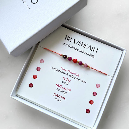 A white gift box that contains a bracelet made of tangerine silk string with five mineral beads, put on a promotional card. The bracelet's theme is Courage, the name of the bracelet is Braveheart which is written in the promotional card. The card also explains each stone and what each stone represents: Red Coral for Courage, Ruby for Valor, Garnet for Force, Pink Tourmaline for Confidence & Self-assertion