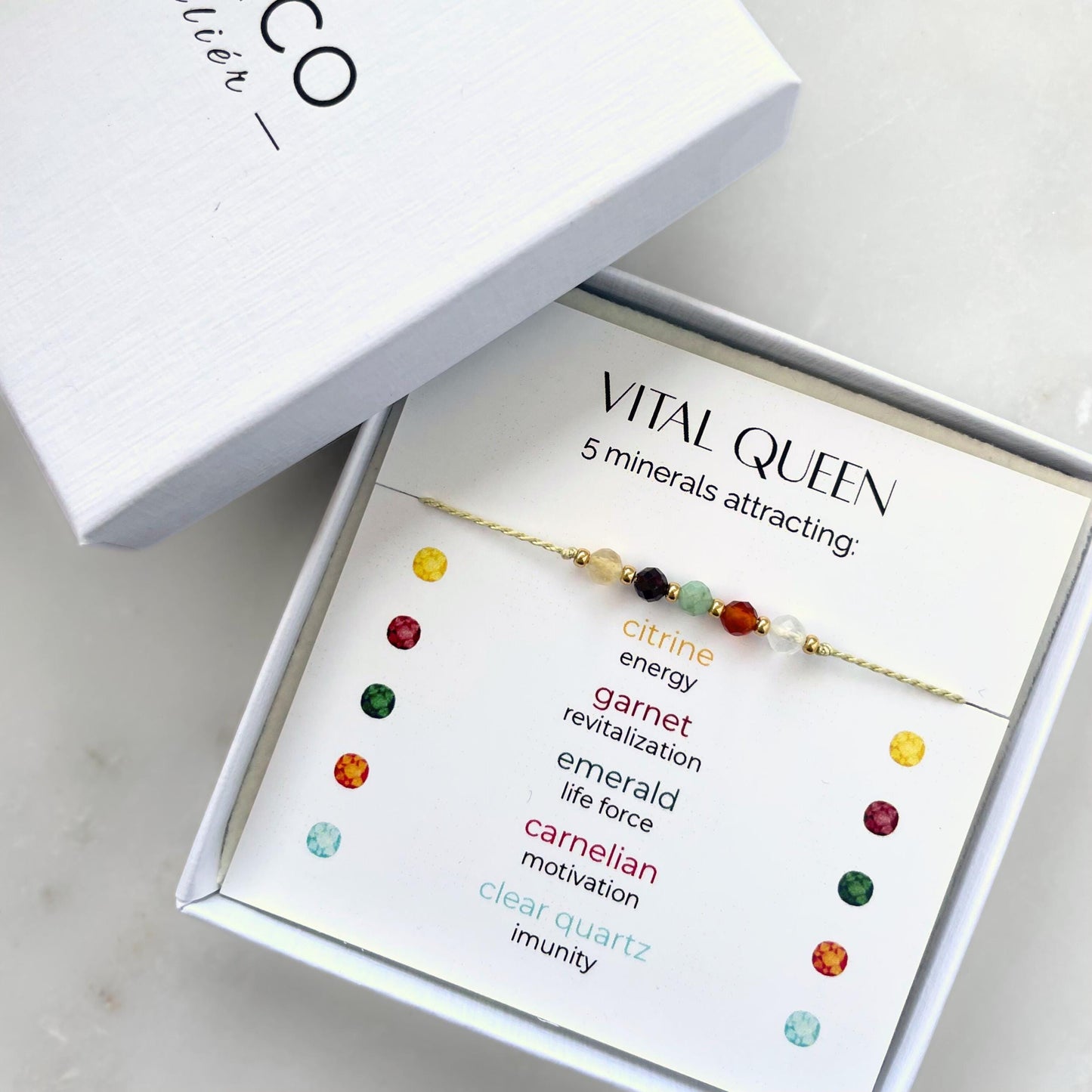 A white gift box that contains a bracelet made of green silk string with five mineral beads, put on a promotional card. The bracelet's theme is Vitality, the name of the bracelet is Vital Queen, which is written in the promotional card. The card also explains each stone and what each stone represents: Citrine for Energy, Garnet for Revitalization, Emerald for Life Force, Carnelian for Motivation, Clear Quartz for Imunity