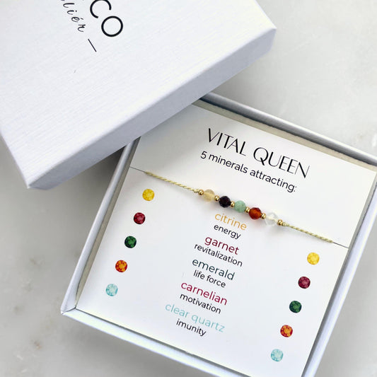 A white gift box that contains a bracelet made of green silk string with five mineral beads, put on a promotional card. The bracelet's theme is Vitality, the name of the bracelet is Vital Queen, which is written in the promotional card. The card also explains each stone and what each stone represents: Citrine for Energy, Garnet for Revitalization, Emerald for Life Force, Carnelian for Motivation, Clear Quartz for Imunity
