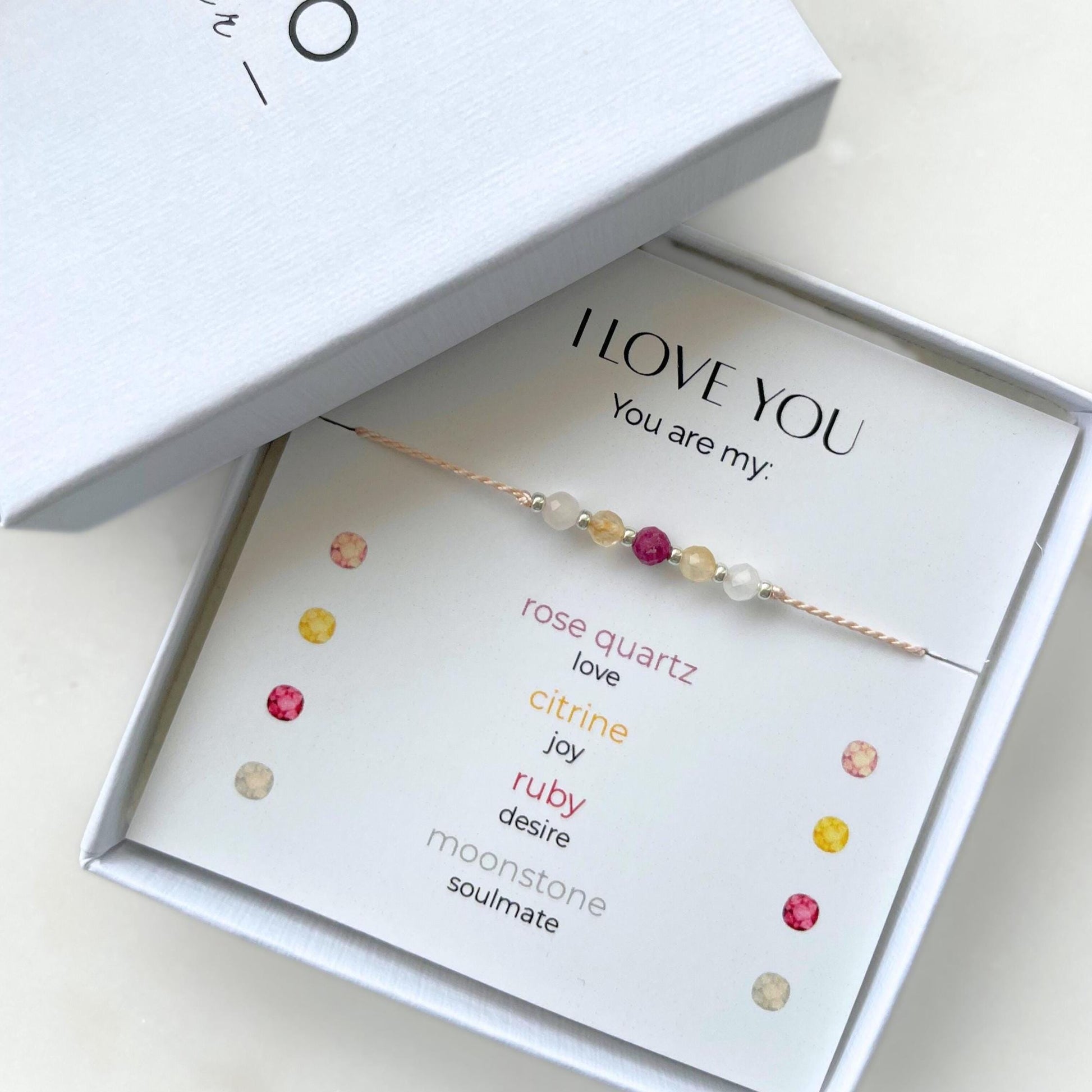 A white gift box that contains a bracelet made of pink silk string with five mineral beads, put on a promotional card. The bracelet's theme is "Love Declaration", the name of the bracelet is "I Love You", which is written in the promotional card. The card also explains each stone and what message each stone sends to the recipient, saying "Your are my": Rose Quartz for Love, Citrine for Joy, Ruby for Desire and Moonstone for Soulmate.
