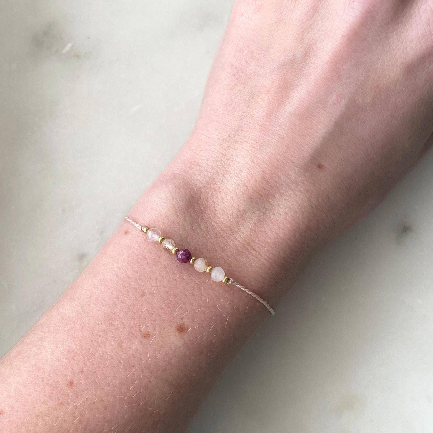 A female hand modeling a pink silk cord bracelet strung with 5 mineral beads: rose quartz, citrine, ruby, another citrine and moonstone, with extra small gold beads between each mineral. 