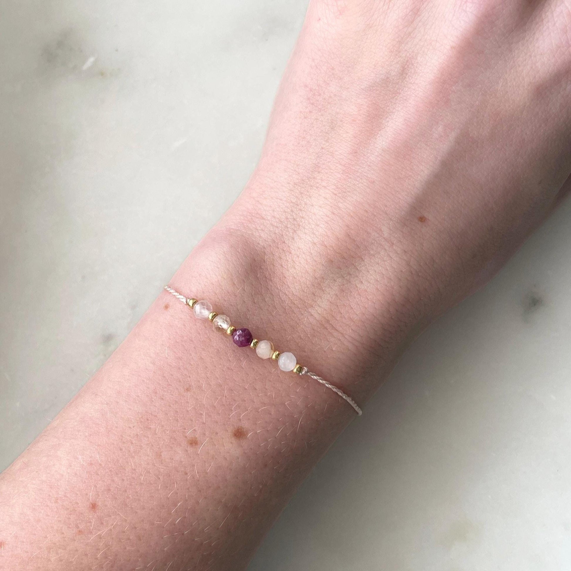 A female hand modeling a pink silk cord bracelet strung with 5 mineral beads: rose quartz, citrine, ruby, another citrine and moonstone, with extra small gold beads between each mineral. 
