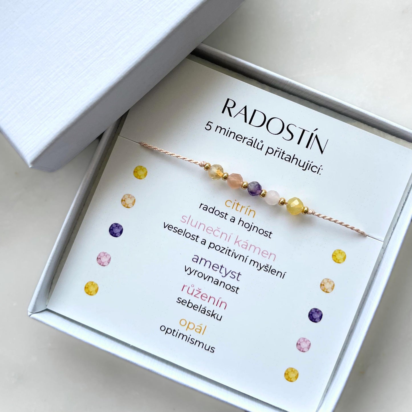 Náramek Radostín z 5 přírodních minerálů v dárkové krabičce. Karta v balení popisuje symboliku kamenů: Citrín pro radost a hojnost, sluneční kámen pro veselost a pozitivitu, ametyst pro vyrovnanost, růženín pro sebelásku, opál pro optimismus