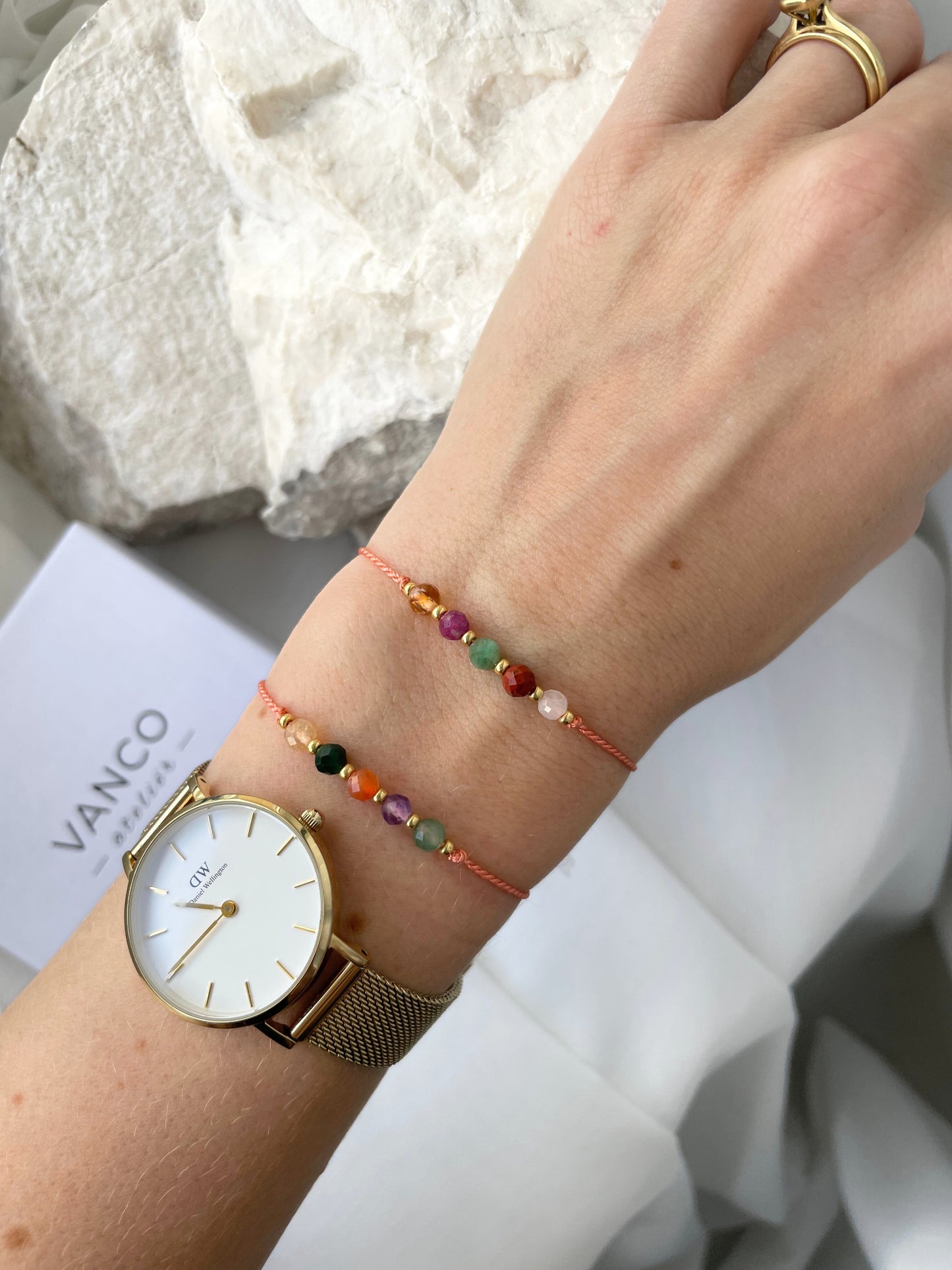 A female hand modeling a tangerine color silk cord bracelet strung with five mineral beads: white moonstone, red jasper, green emerald, pink ruby, yellow citrine. Each mineral is separated from another with a little gold bead, making the design more elegant. Model is also wearing a classic DW round watch with gold metal strap, showing how the bracelet can be a lovely addition to wear and layer with a watch. 