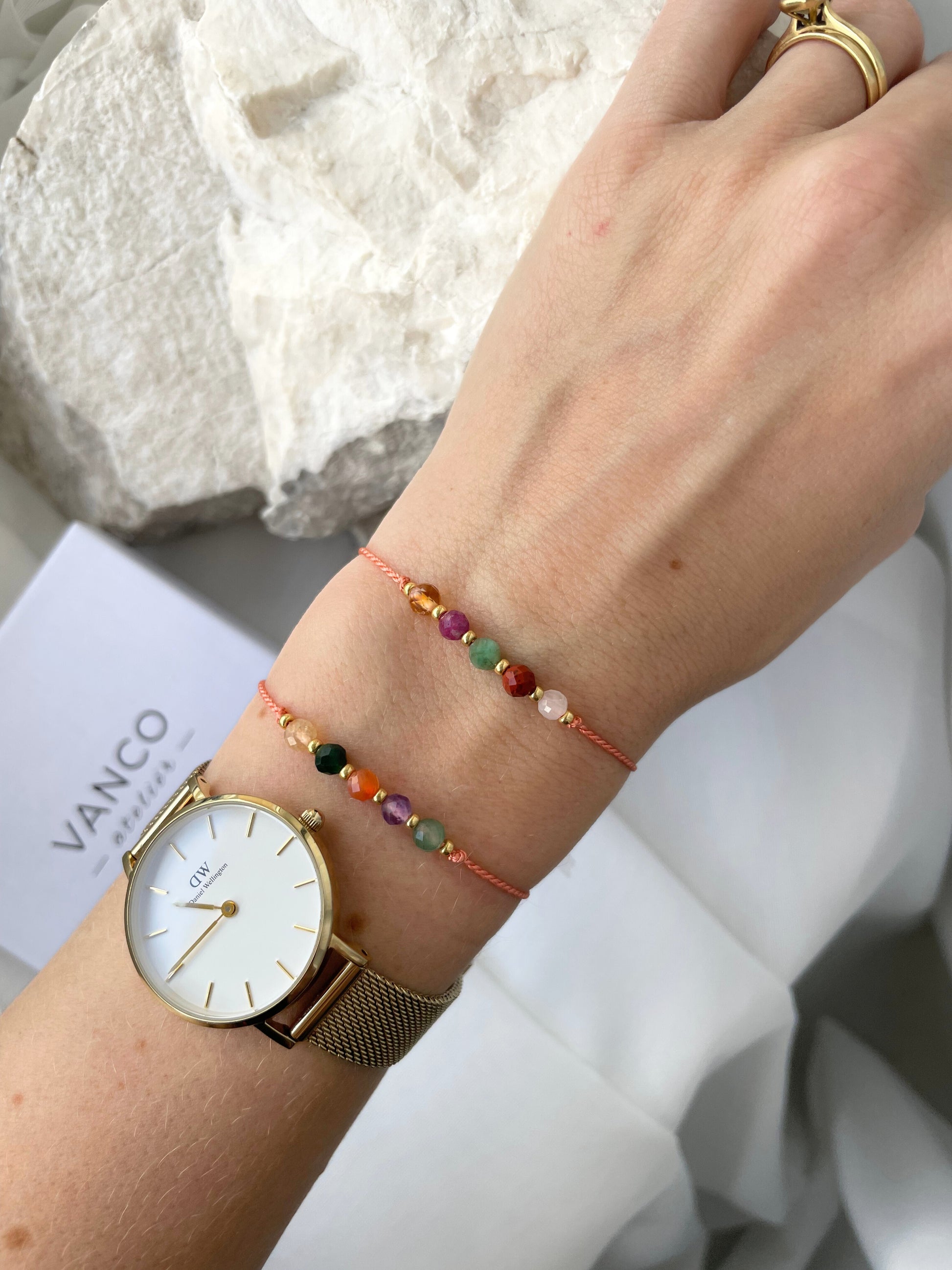 A female hand modeling a tangerine color silk cord bracelet strung with five mineral beads: white moonstone, red jasper, green emerald, pink ruby, yellow citrine. Each mineral is separated from another with a little gold bead, making the design more elegant. Model is also wearing a classic DW round watch with gold metal strap, showing how the bracelet can be a lovely addition to wear and layer with a watch. 