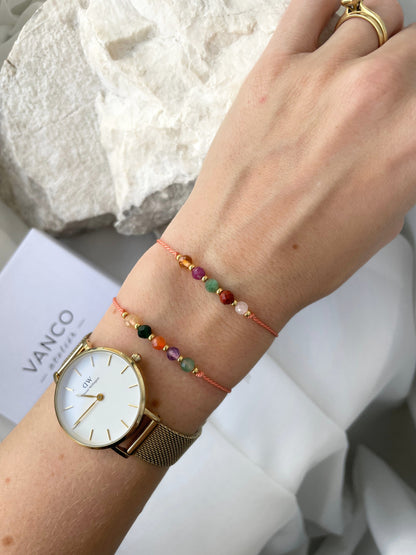 A female hand modeling a tangerine color silk cord bracelet strung with five mineral beads: white moonstone, red jasper, green emerald, pink ruby, yellow citrine. Each mineral is separated from another with a little gold bead, making the design more elegant. Model is also wearing a classic DW round watch with gold metal strap, showing how the bracelet can be a lovely addition to wear and layer with a watch. 